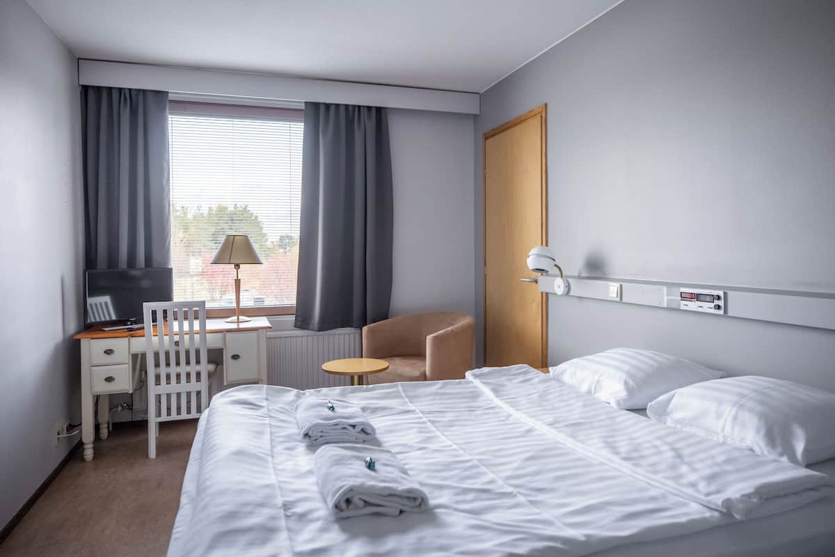 Une chambre d’hôtel avec un lit, un bureau, une chaise, une lampe et une fenêtre avec des rideaux.