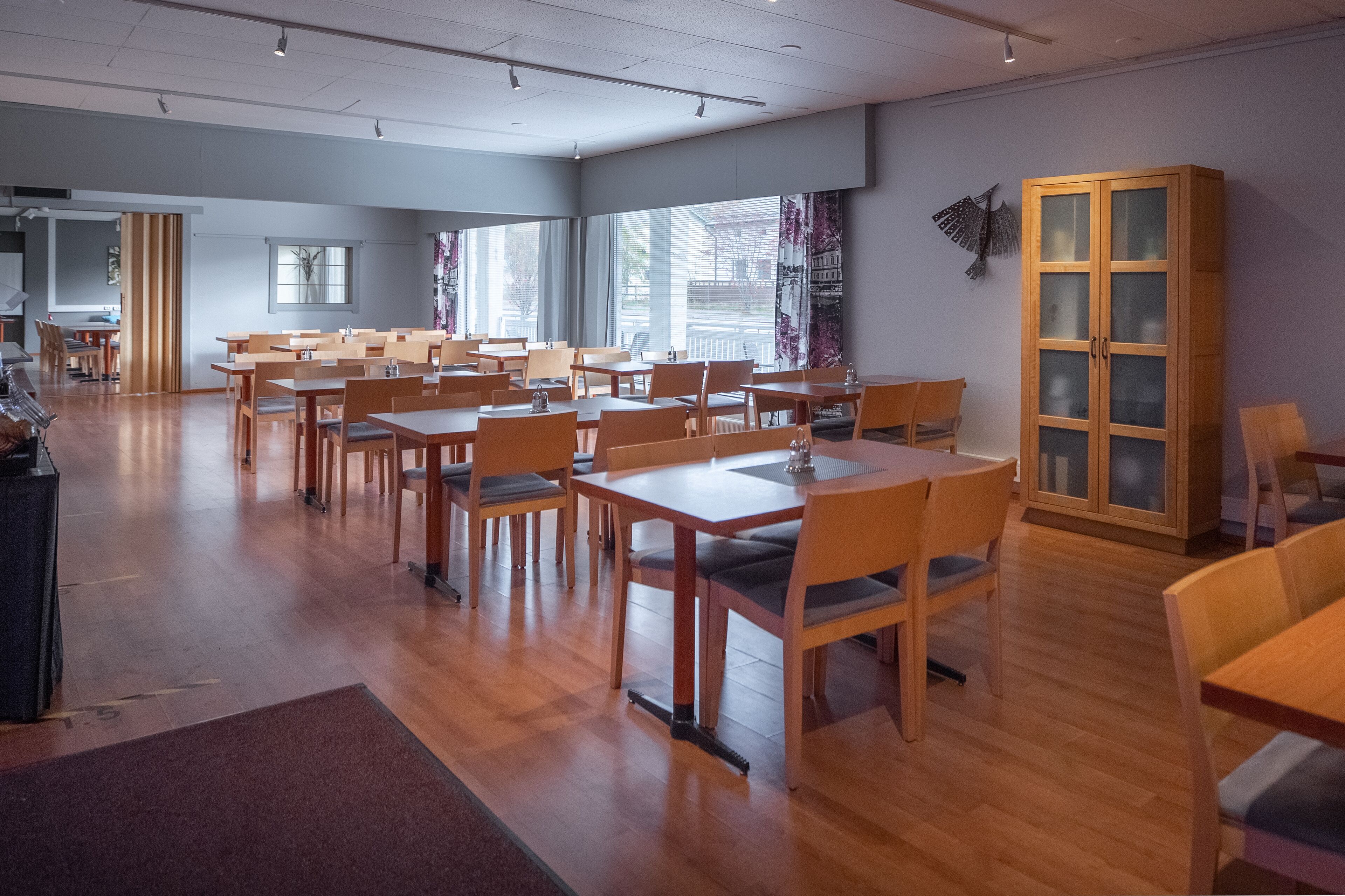 A spacious dining area with wooden tables and chairs, large windows, and a cabinet with glass doors.