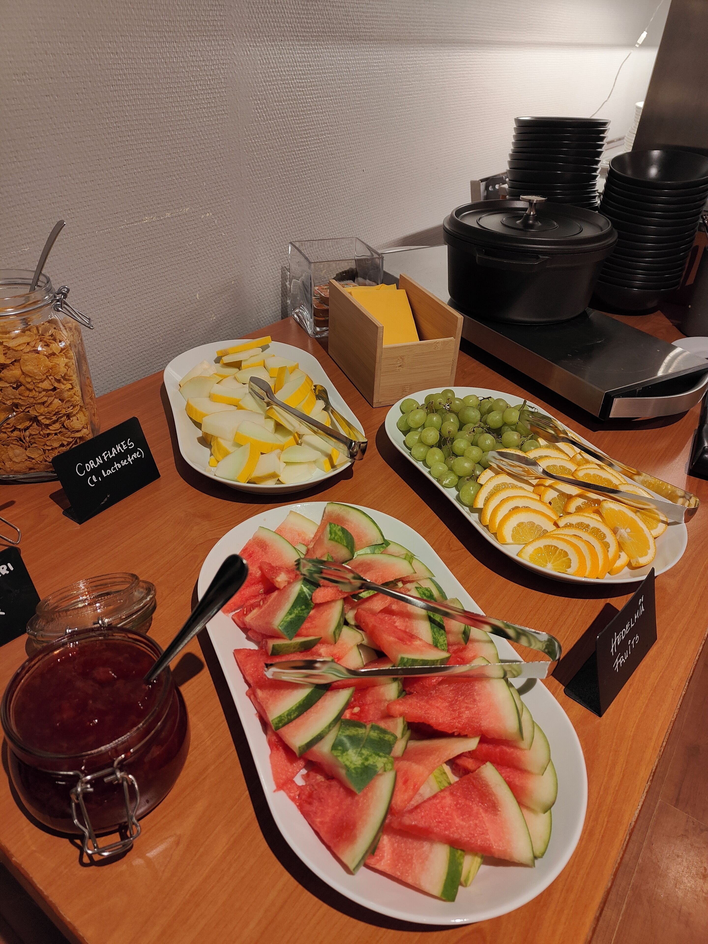 Ein Buffet mit verschiedenen Speisen, darunter Cornflakes, Apfelscheiben, Trauben, Wassermelone und Orangenscheiben.
