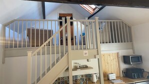 Une chambre mansardée avec un plafond en pente, un escalier blanc avec une rampe, une poutre en bois au plafond, un puits de lumière, un lit, un petit coin cuisine avec un four à micro-ondes, un grille-pain et une machine à café.