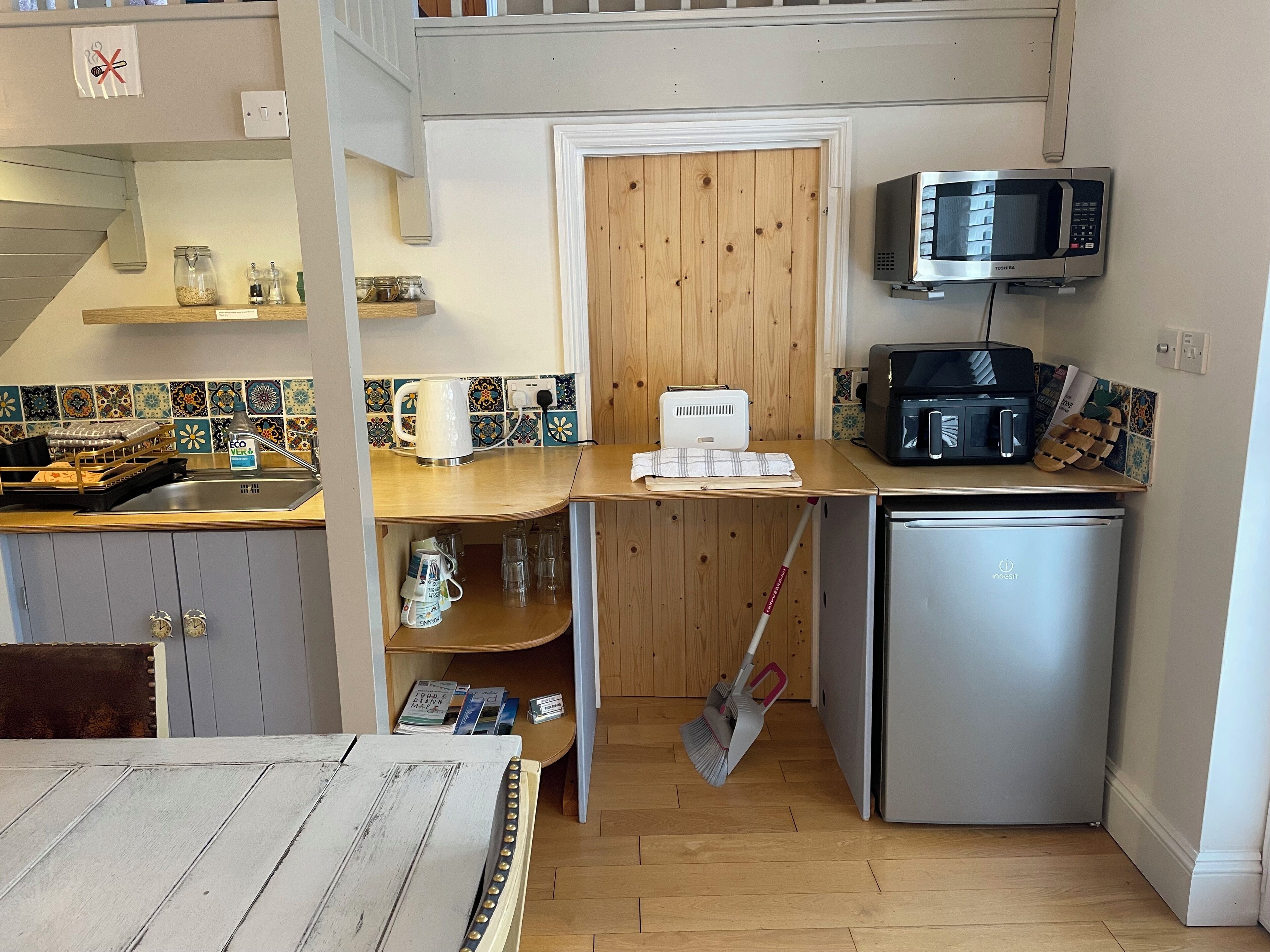 A compact kitchen with a microwave, toaster, and refrigerator. There is a wooden door leading to another room, and a broom leaning against the wall.