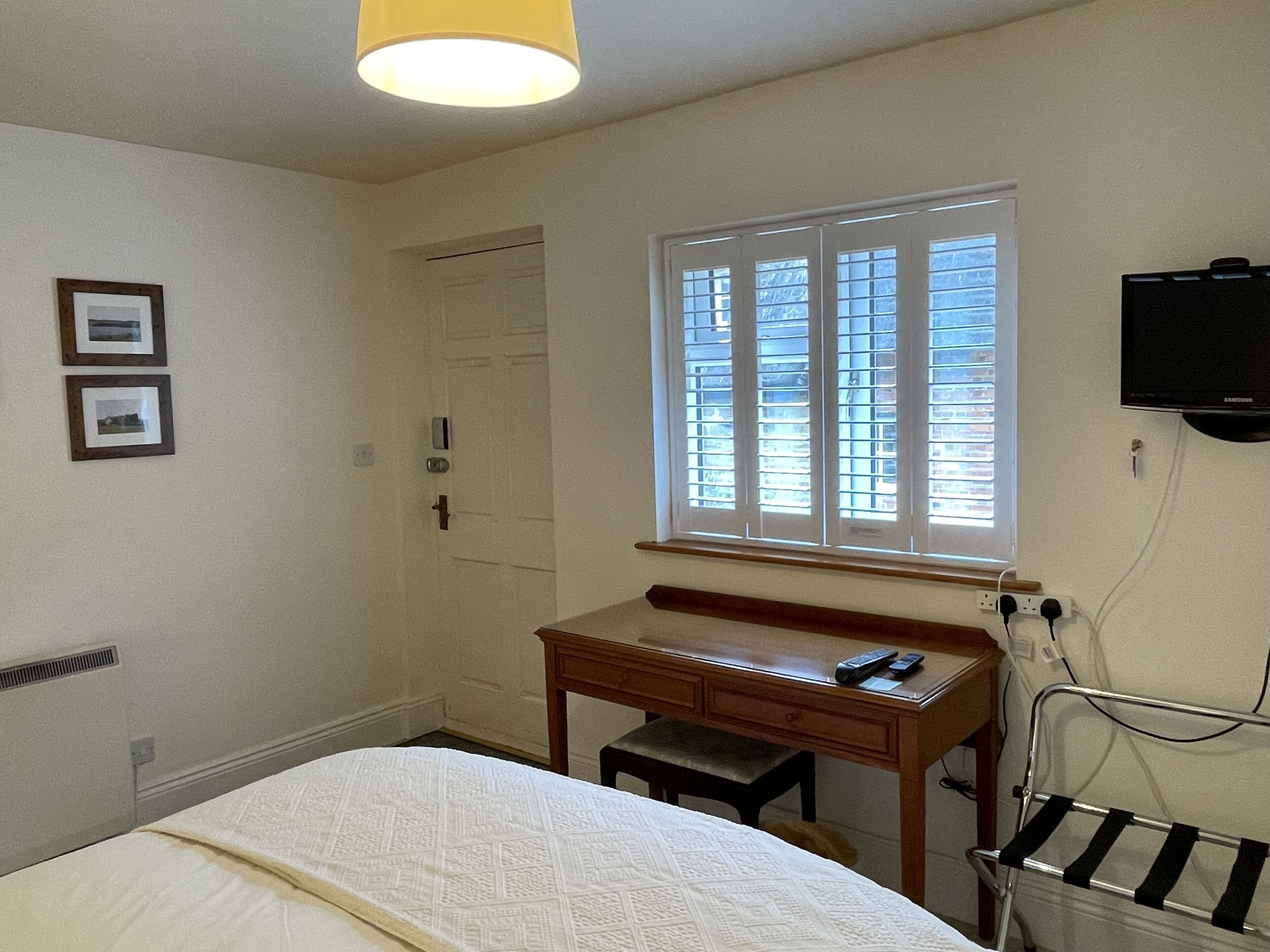A bedroom with a bed, a desk, a chair, a window with blinds, a TV mounted on the wall, and a radiator.