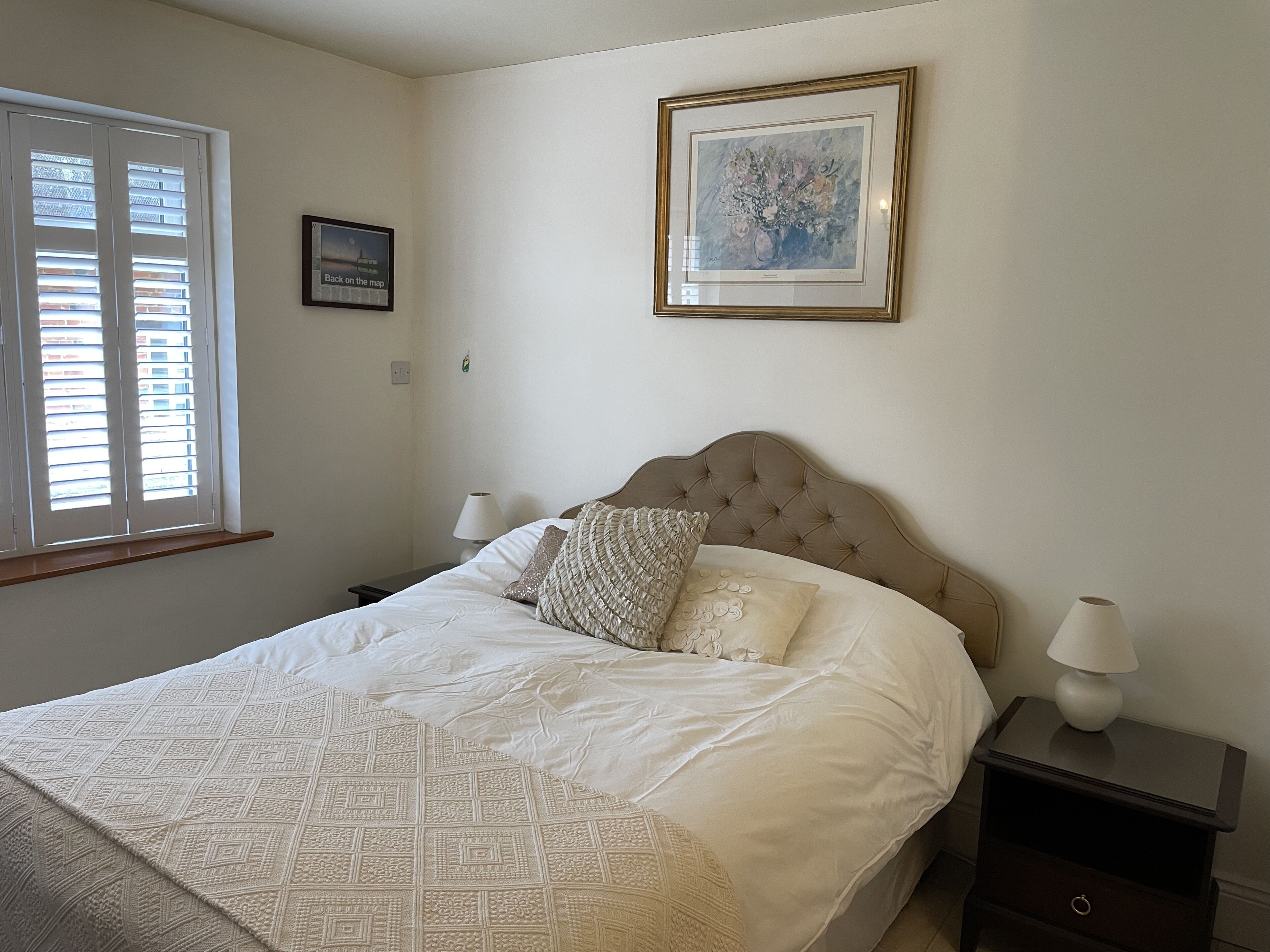 A neatly made bed with a patterned bedspread, a framed artwork above it, and a window with blinds on the left.