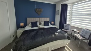 A bedroom with a bed, bedside tables, a chair, a heart-shaped wall decoration, and a window with blinds. - The Clifton at Paignton (Paignton)