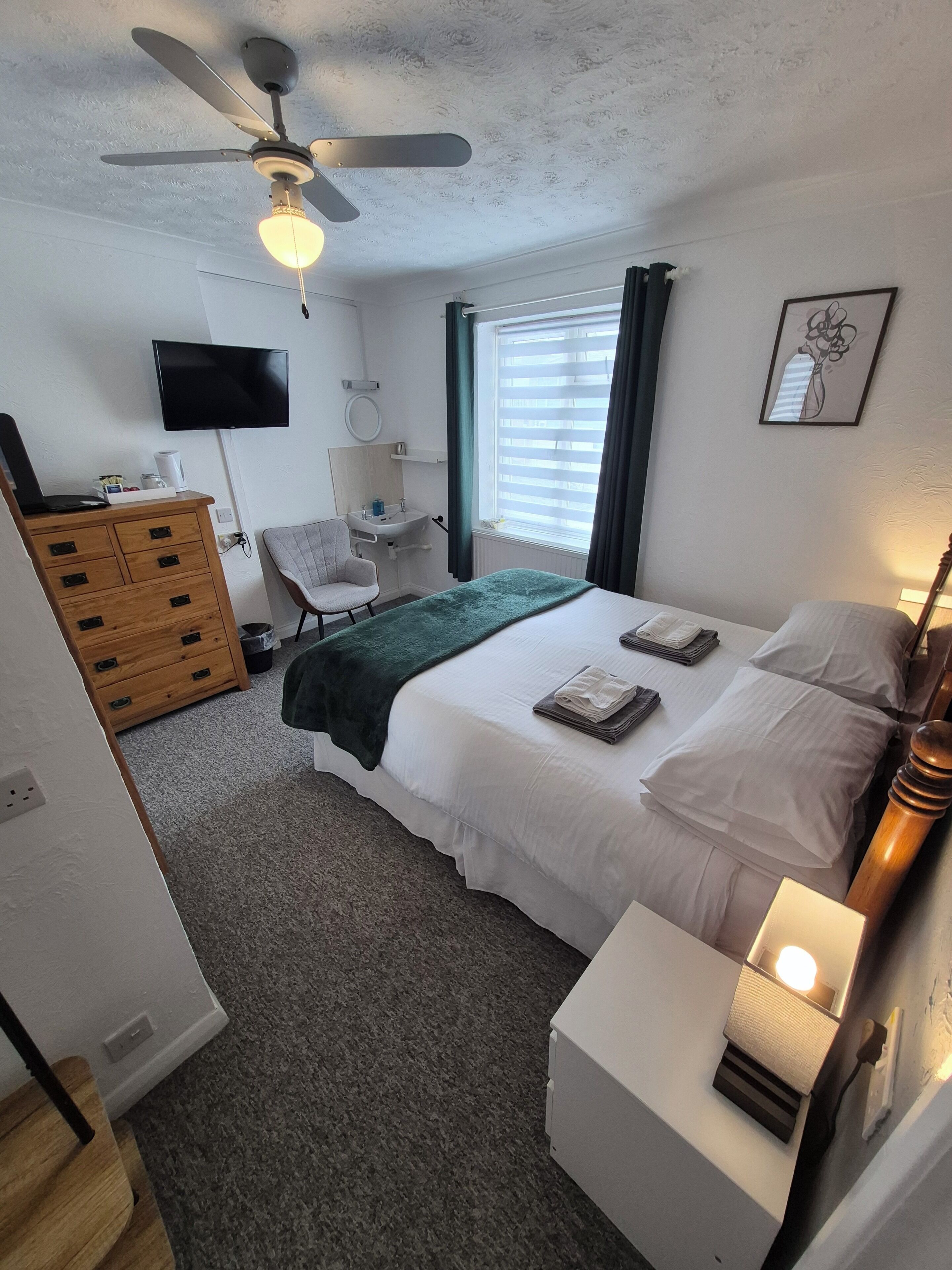 A bedroom with a bed, bedside table, lamp, ceiling fan, TV, and a desk with a chair.