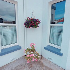 Um canto com duas janelas, um cesto de flores pendurado e um vaso com flores cor-de-rosa.