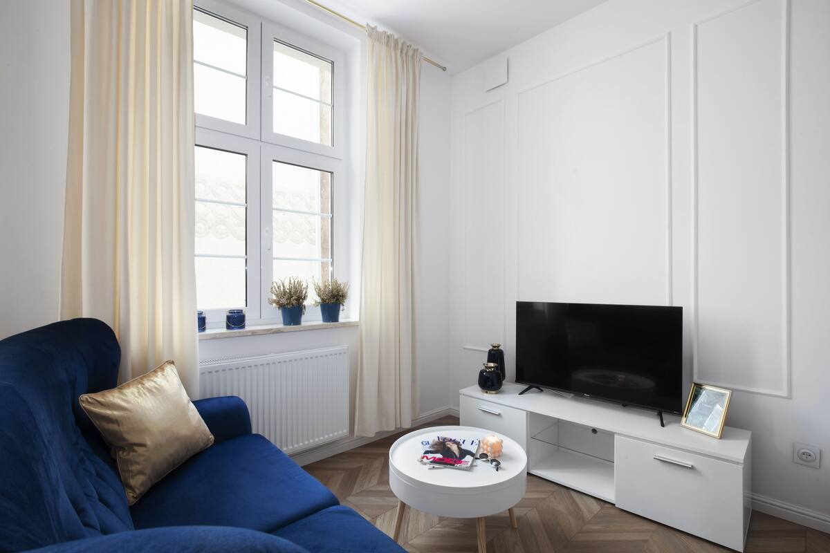 A modern living room with a blue sofa, a white coffee table, a flat-screen TV, and a window with curtains.