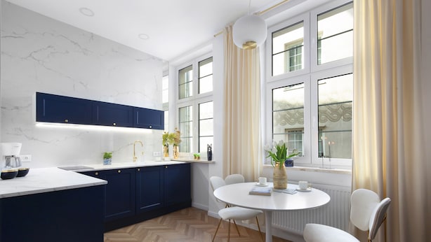 Una cocina moderna con armarios azul oscuro, encimera blanca y suelo de madera en patrón de espiga. Hay una mesa redonda con sillas blancas y una lámpara colgante blanca encima.