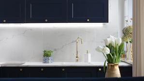 Una cocina con armarios oscuros, encimera blanca, grifo dorado y un florero con flores blancas.