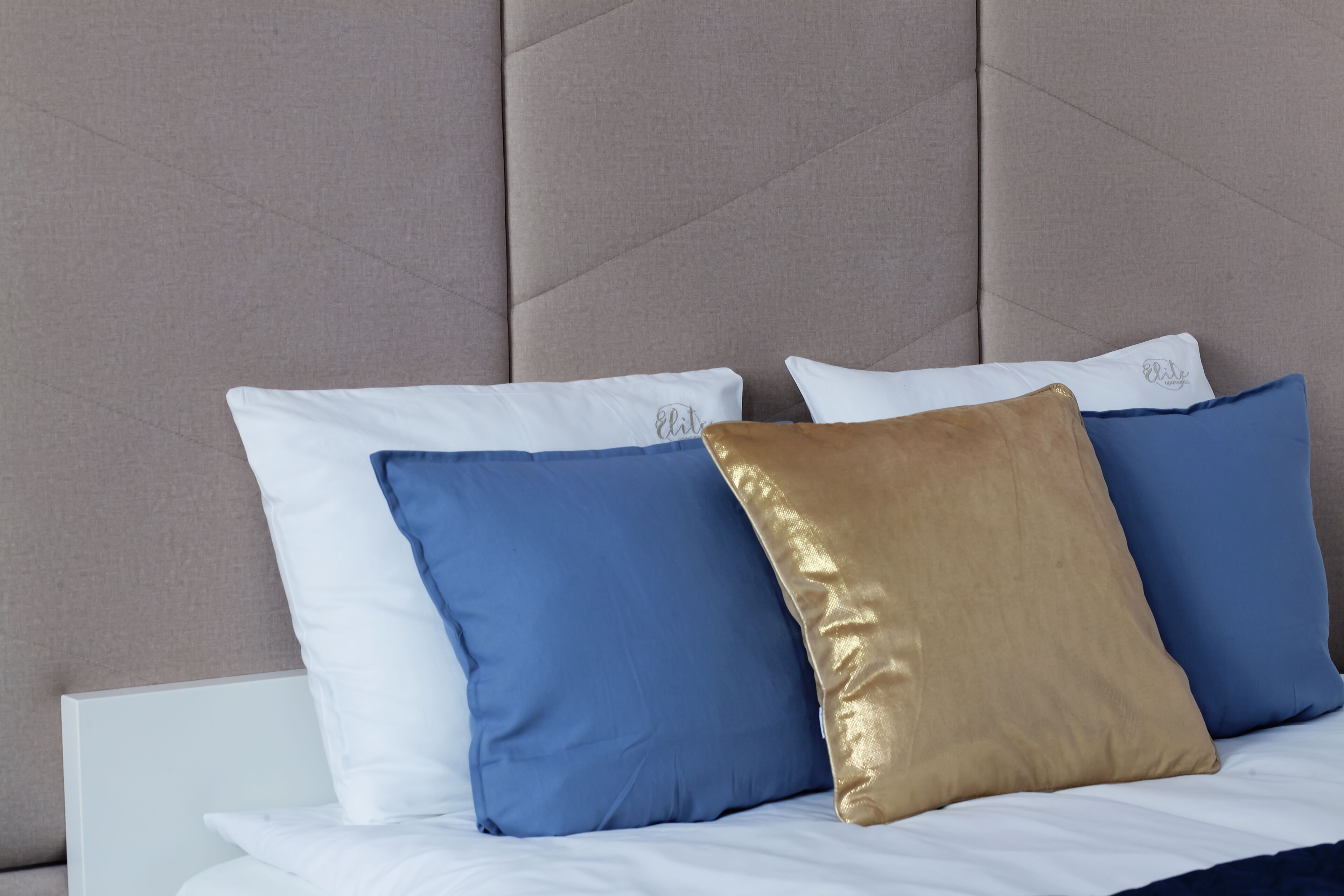 A bed with white bedding and three pillows in blue, gold, and white.