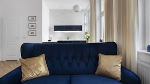 Una sala de estar moderna con un sofá azul oscuro, dos cojines dorados, una pared blanca con marcos y cuadros, una cocina al fondo y una ventana con cortinas.