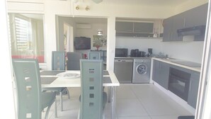 A compact kitchen with a washing machine, a dining area with chairs, and a TV in the background. - Beachfront the dream holiday (saint martin)