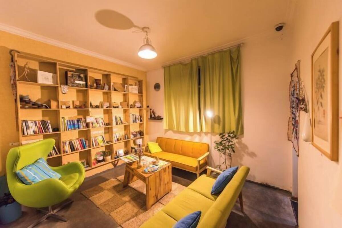 A cozy living room with a bookshelf, a green armchair, a yellow sofa, and a coffee table.