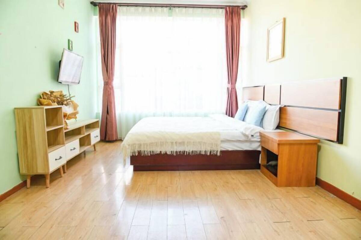 A bedroom with a bed, wooden nightstand, built-in shelving, a TV, and a window with curtains.