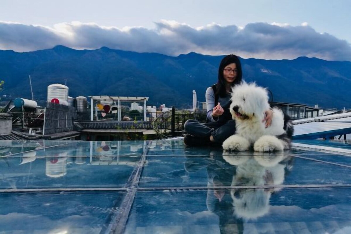 Une personne assise sur un plancher de verre, avec un chien, des montagnes en arrière-plan et des structures industrielles.