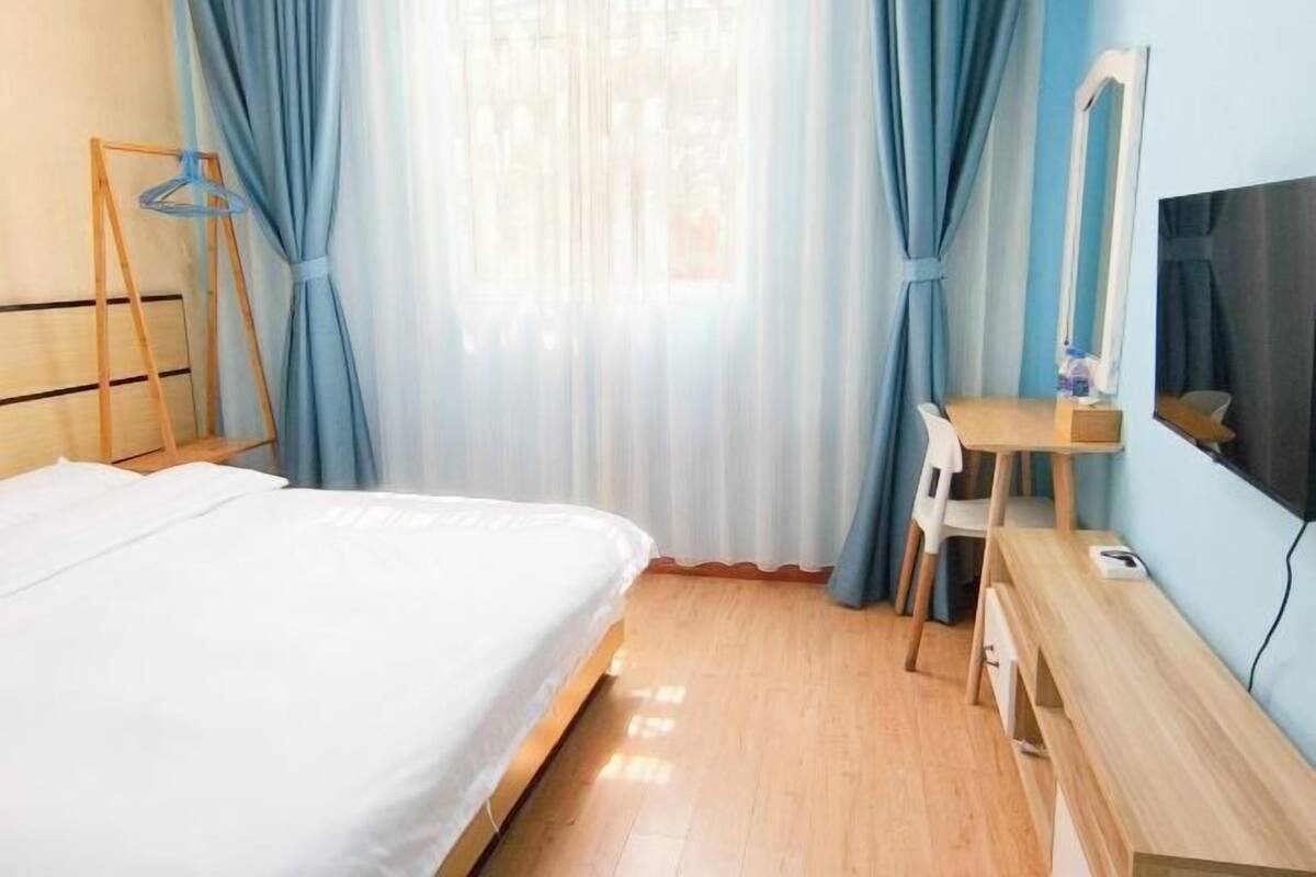 A hotel room with a bed, a wooden desk, a chair, a mirror, and a window with curtains.