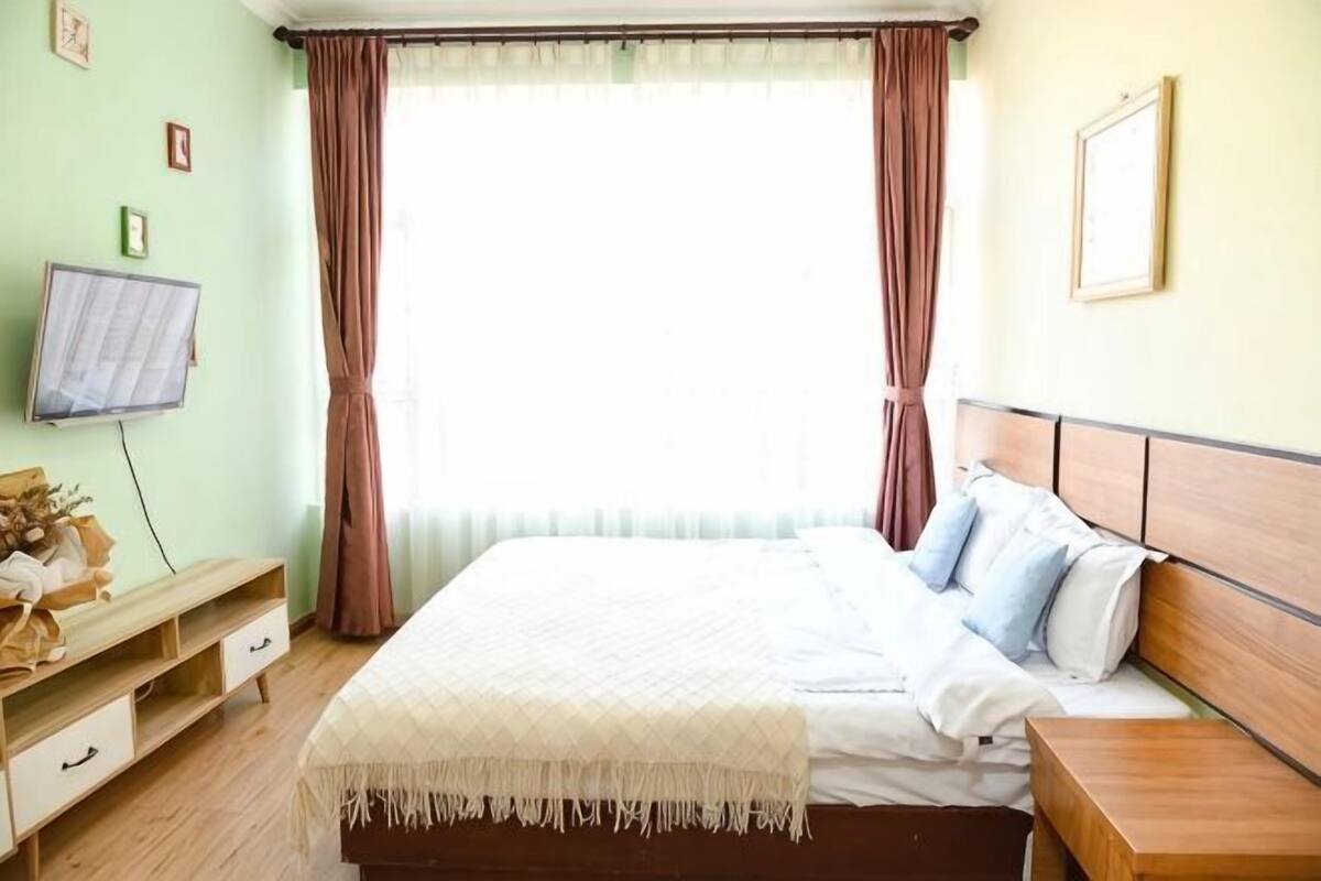 A bedroom with a bed, bedside table, wooden headboard, and a window with curtains.