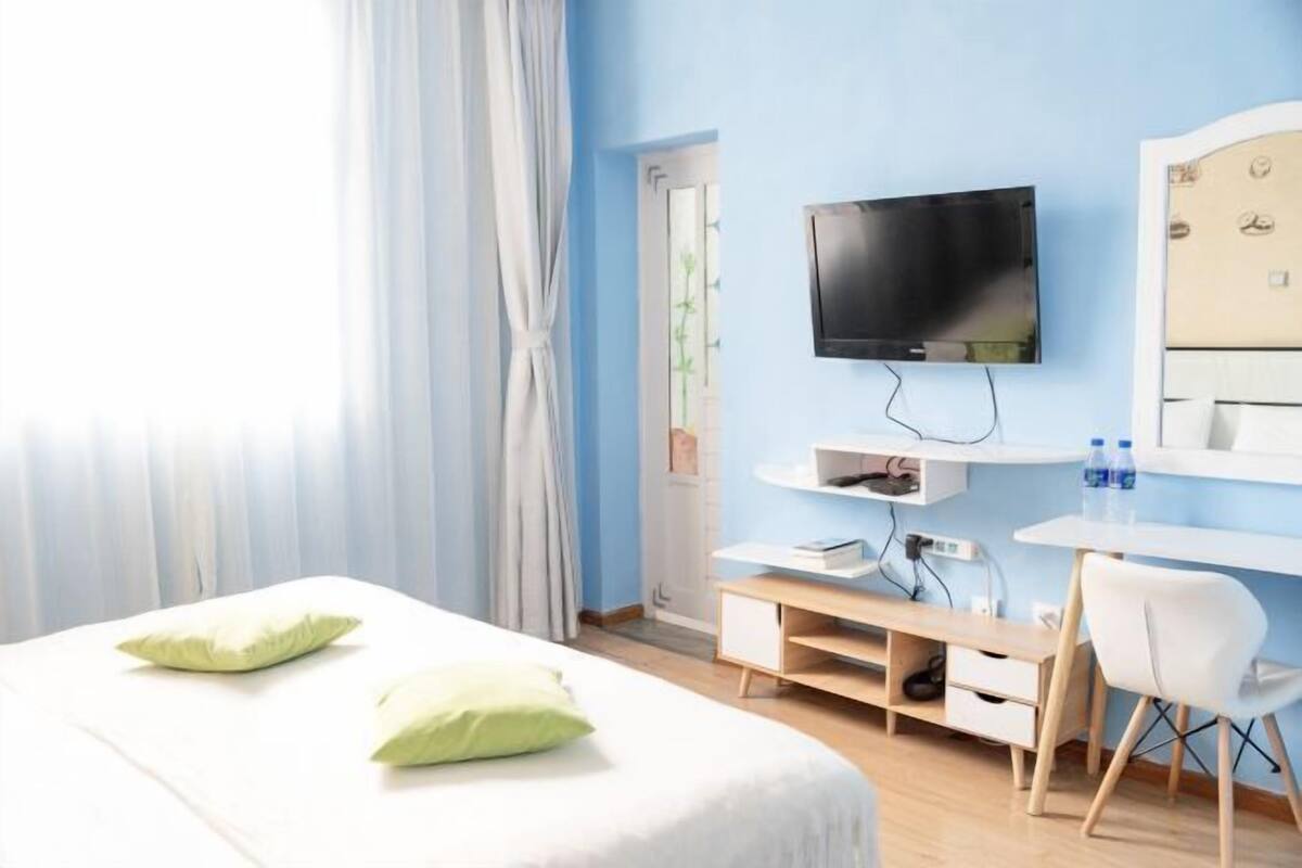 A hotel room with a bed, a TV mounted on the wall, a desk with a chair, and a window with curtains.