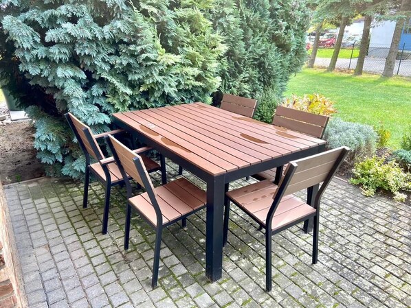 Eine Terrasse im Freien mit einem Holztisch und -stühlen, umgeben von viel Grün und einem gepflasterten Weg.