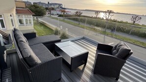 Eine Terrasse mit Korbmöbeln und einem Glasgeländer mit Blick auf ein Gewässer und eine Straße.