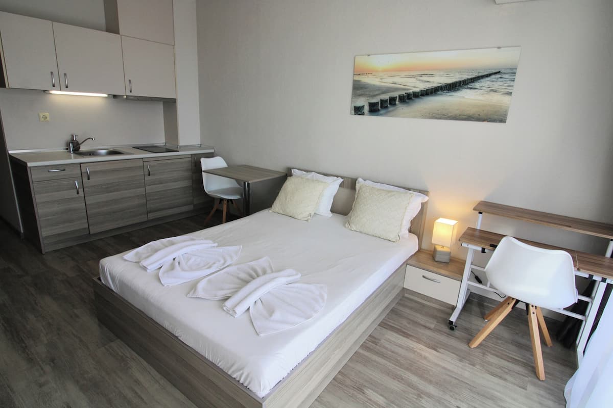 A modern hotel room with a large bed, a desk, a chair, a small table, and a painting of a beach scene.
