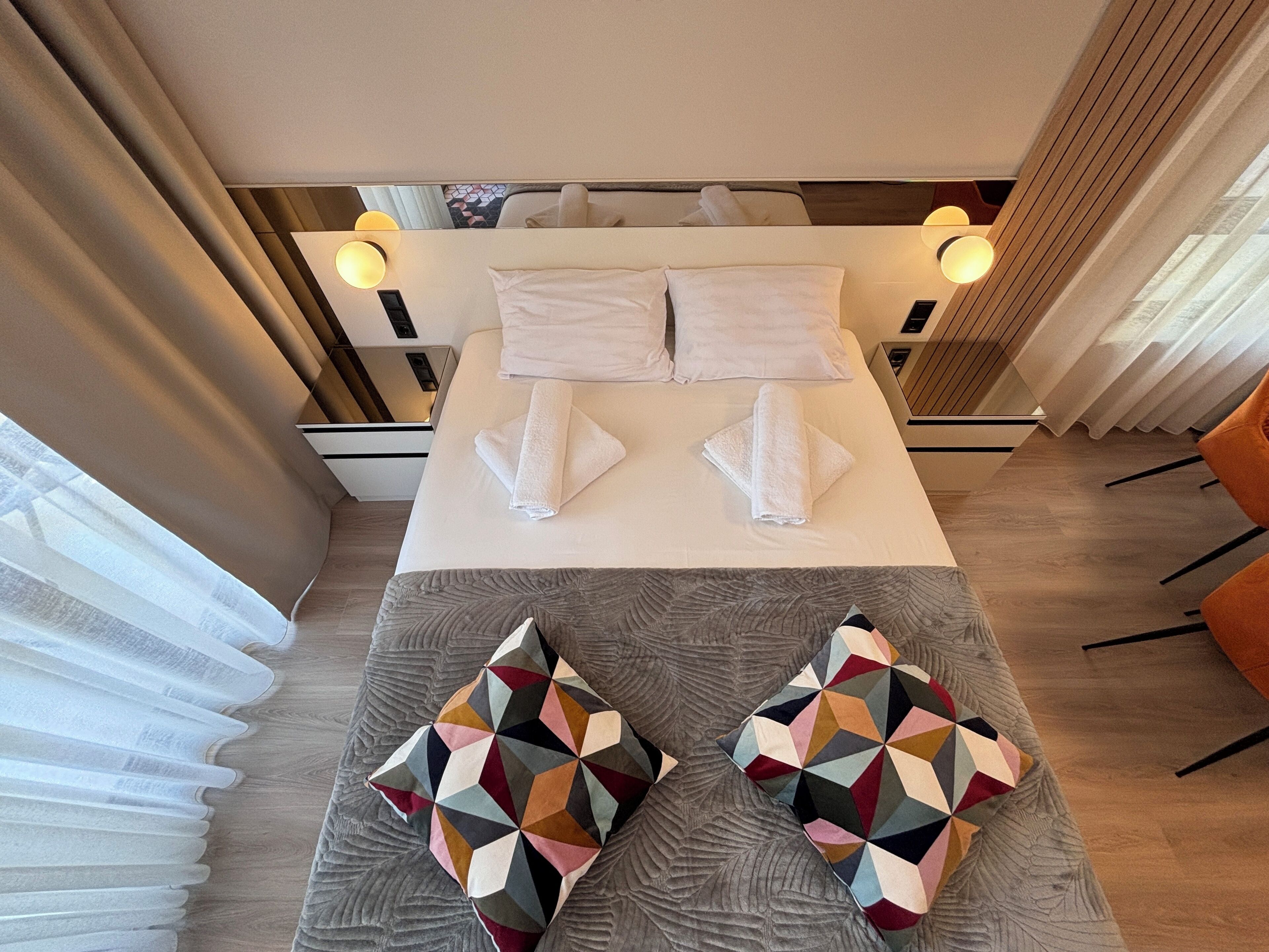 A hotel room with a bed, two pillows, a bedside table, and a chair.