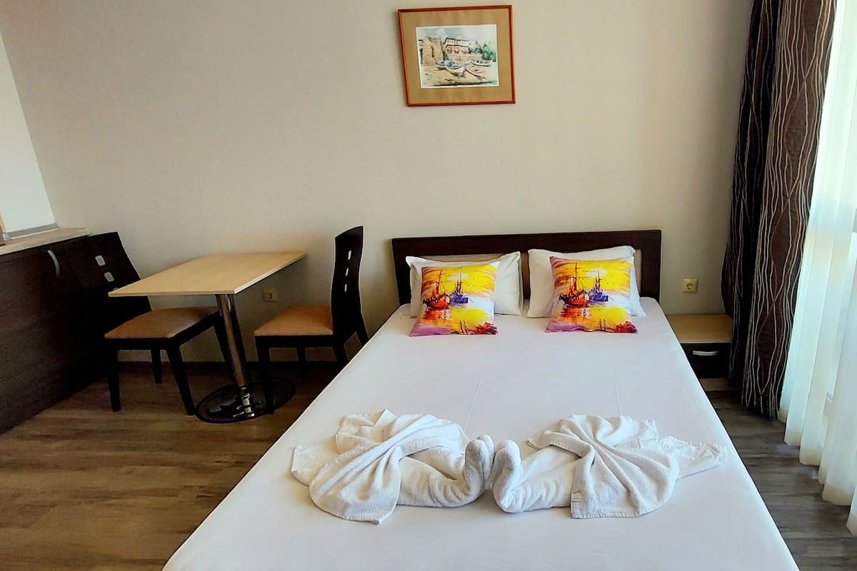 A hotel room with a bed, two pillows with a sunset design, a small table, a chair, a cabinet, and a window with curtains.