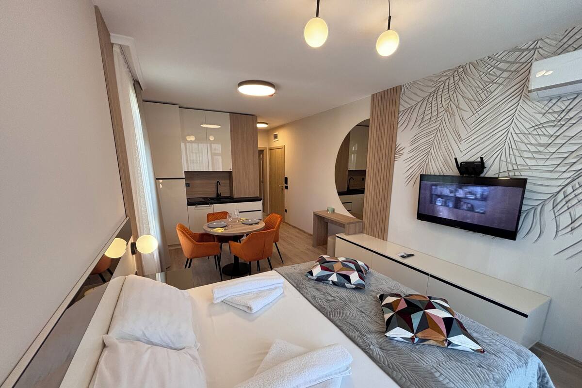 A modern hotel room with a bed, a TV, a dining area, and a kitchen.
