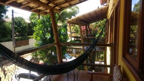 A hammock under a wooden pergola with a view of a garden and a building.