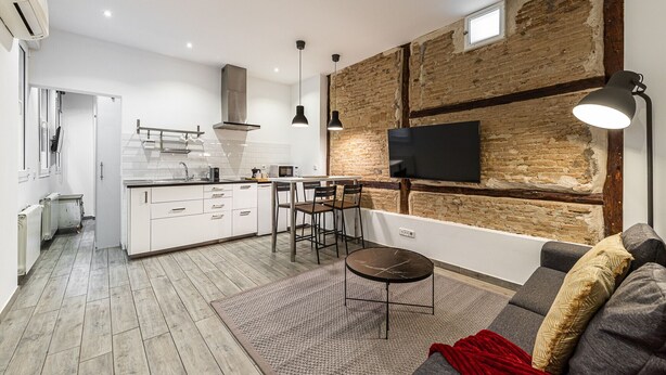 Een moderne keuken met witte kastjes, een zwart aanrechtblad en een ingebouwd fornuis. Er is een zithoek met een ronde tafel en stoelen, en een grote tv aan een bakstenen muur bevestigd.