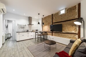 Een moderne keuken met witte kastjes, een zwart aanrechtblad en een ingebouwd fornuis. Er is een zithoek met een ronde tafel en stoelen, en een grote tv aan een bakstenen muur bevestigd.