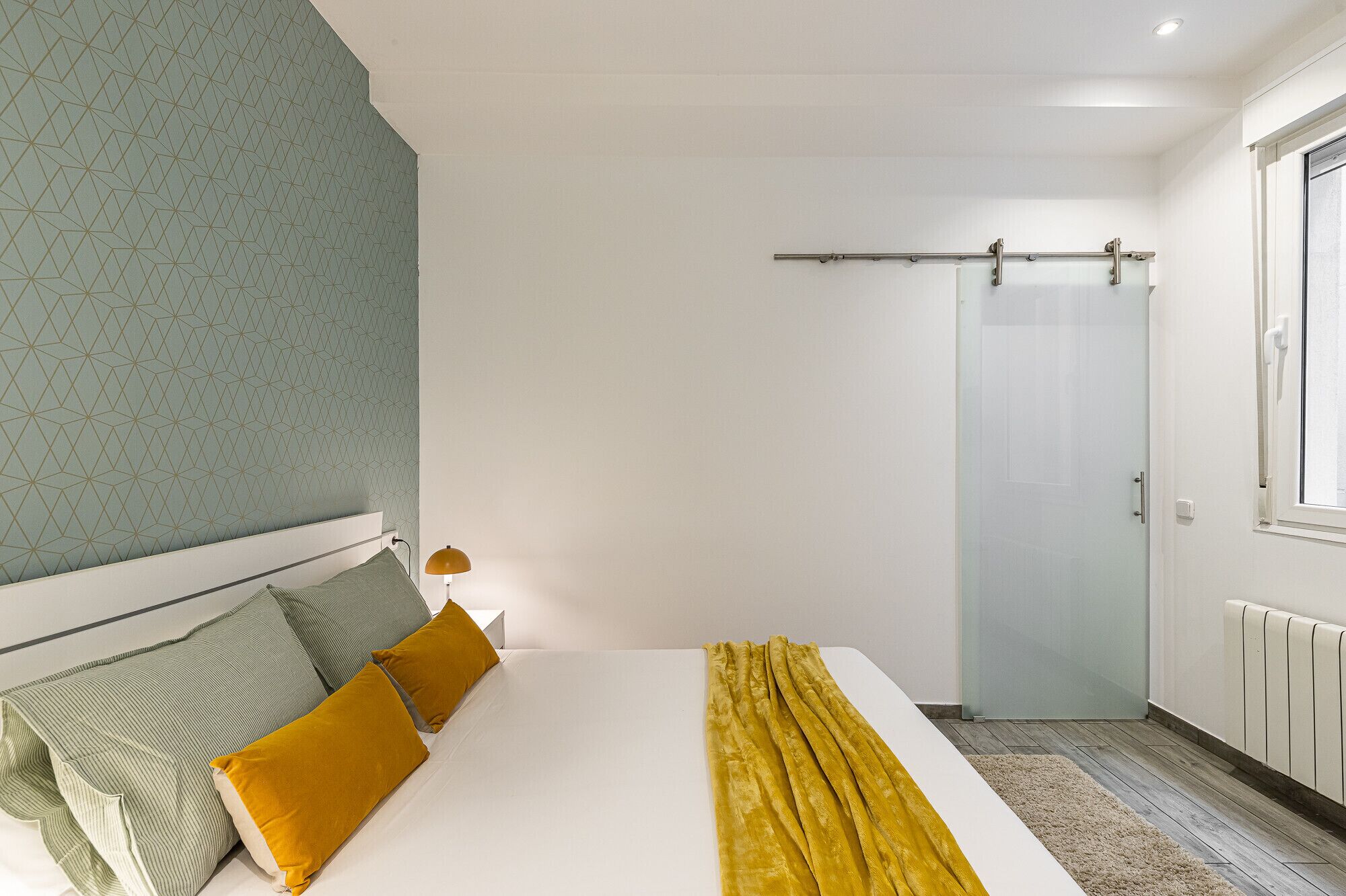A hotel room with a bed, a nightstand, a lamp, a window with a curtain, and a radiator.