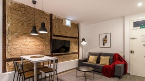 Een moderne woonkamer met een metselwerk accentmuur, een eethoek met een witte tafel en stoelen, een grijze bank met kussens en een rode plaid.