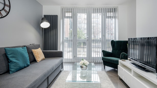 A modern living room with a grey sofa, teal and patterned cushions, a glass coffee table with a vase of white flowers, a dark green armchair, a flat-screen TV on a white stand, and a large window with blinds. - Luxury Chic Apartment near Canary Wharf, Excel, O2 & Stratford (London)