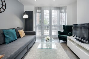 A modern living room with a grey sofa, teal and patterned cushions, a glass coffee table with a vase of white flowers, a dark green armchair, a flat-screen TV on a white stand, and a large window with blinds. - Luxury Chic Apartment near Canary Wharf, Excel, O2 & Stratford (London)