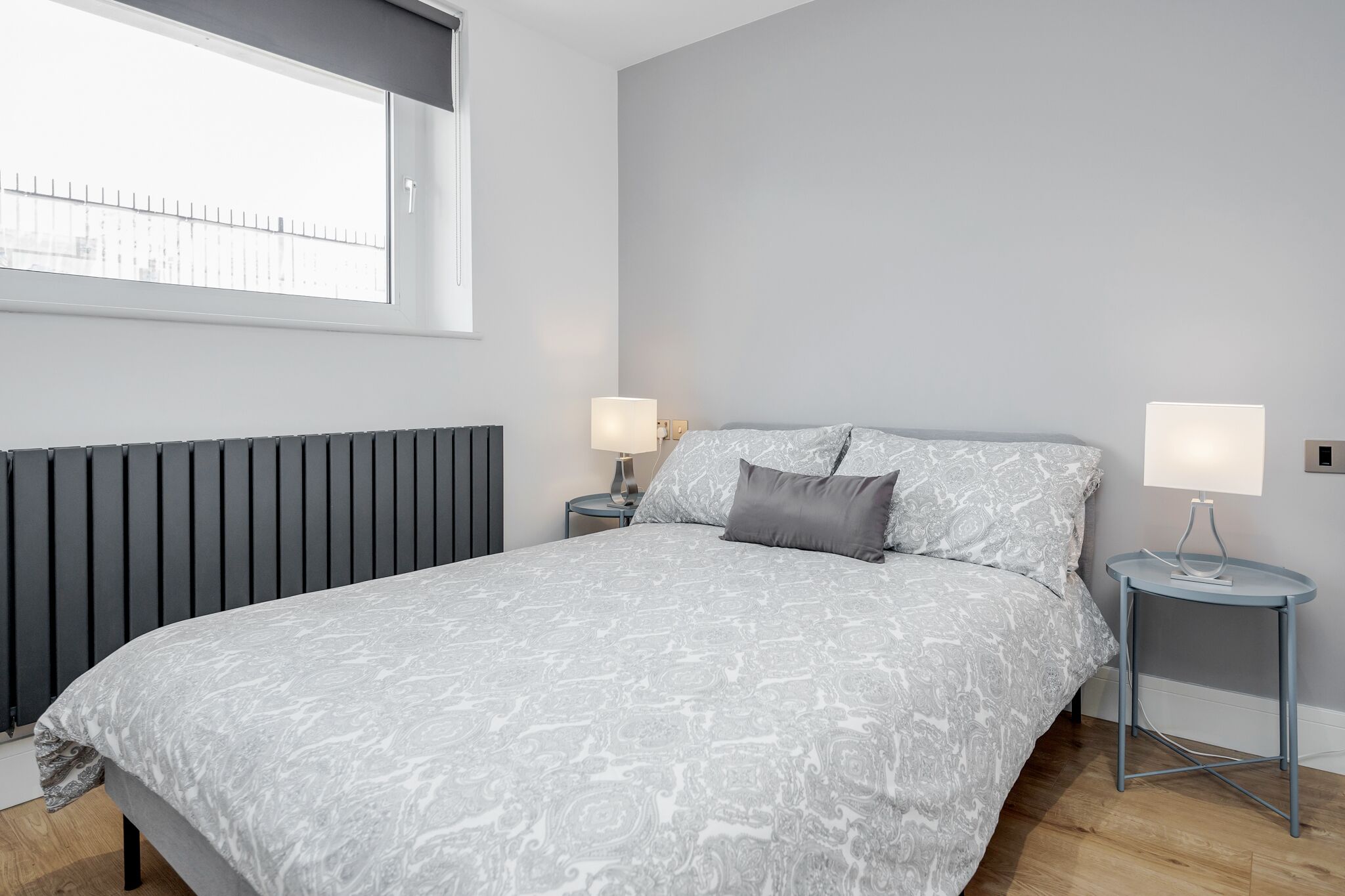 A bedroom with a bed, two bedside tables, a window, and a radiator.