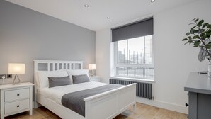 A modern bedroom with a white bed, bedside tables, a radiator, a window with a view of buildings, and a plant. - Luxury Chic Apartment near Canary Wharf, Excel, O2 & Stratford (London)