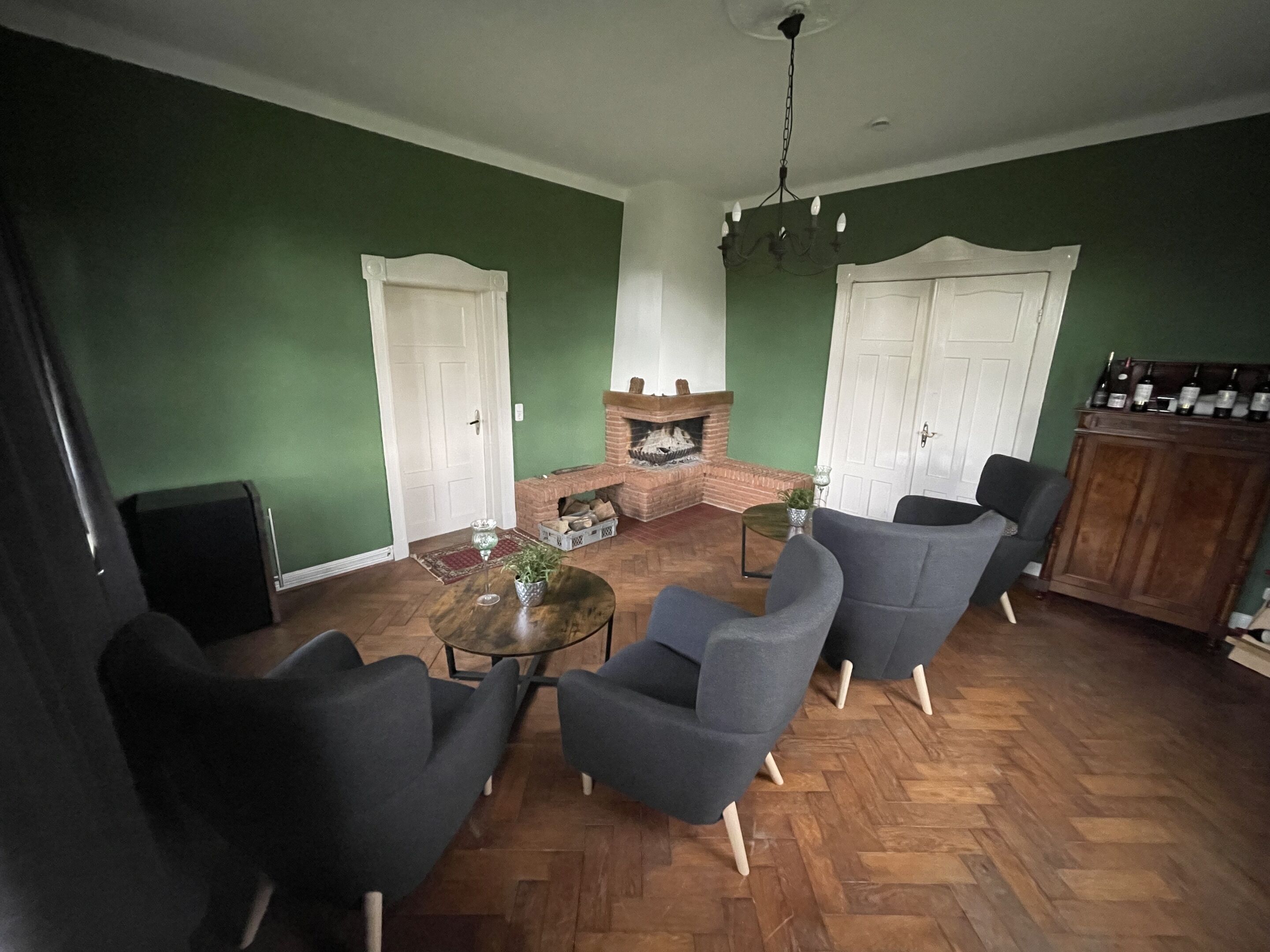 A living room with green walls, a brick fireplace, and a wooden herringbone floor. There are three grey armchairs and a coffee table.