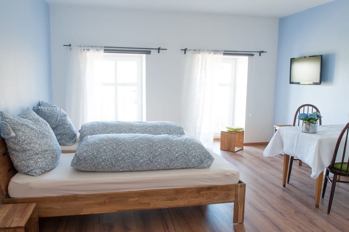 A bedroom with a wooden bed, two windows with curtains, a small table with a plant, and a TV on the wall.