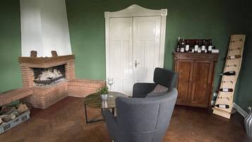 A living room with a brick fireplace, a grey armchair, a small table with a vase, and a wine rack against a green wall.