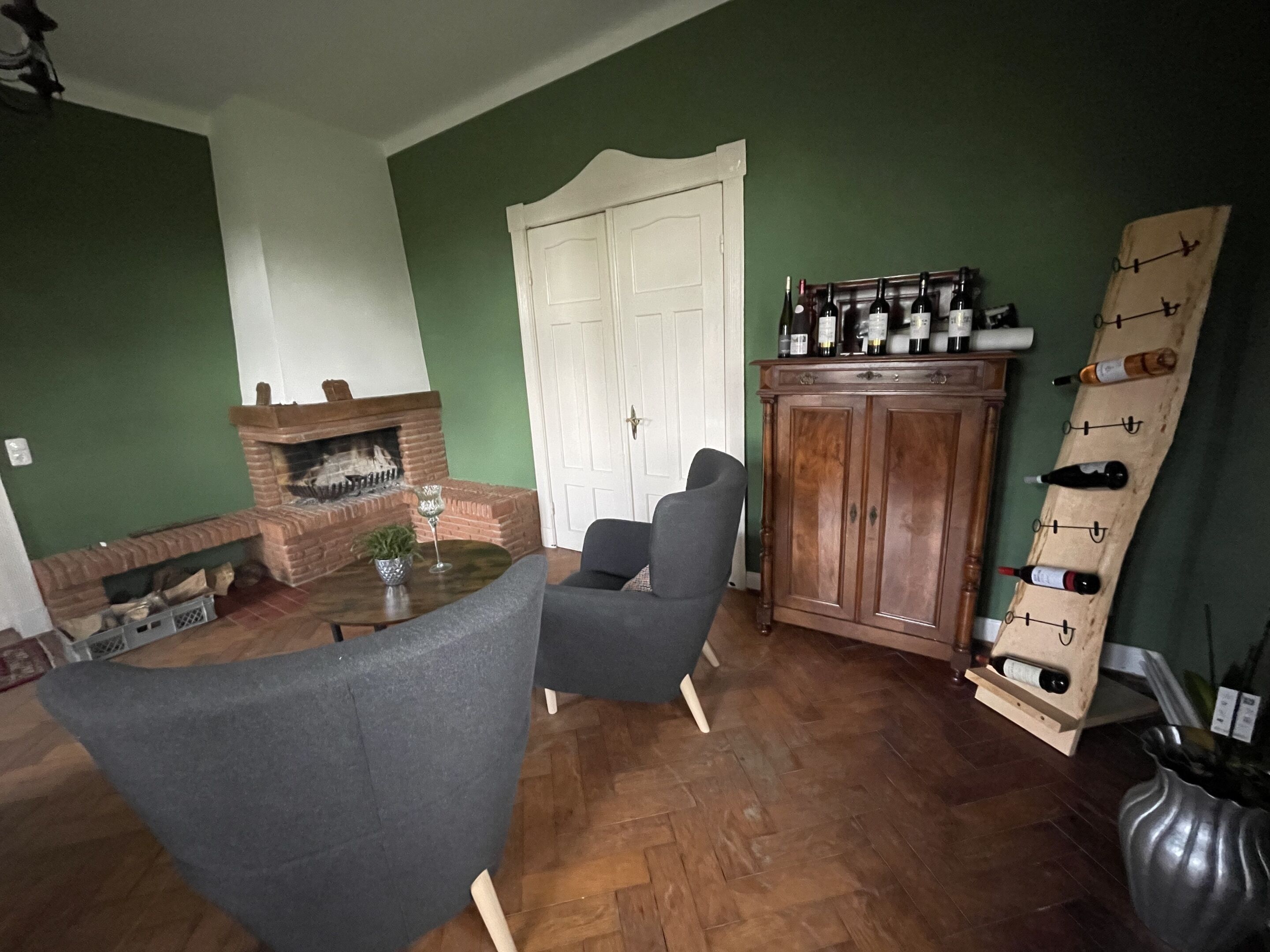 A living room with a brick fireplace, a dark armchair, a wooden cabinet with wine bottles, and a herringbone-patterned wooden floor.