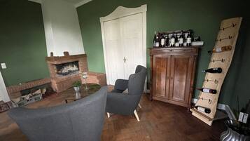 A living room with a brick fireplace, a dark armchair, a wooden cabinet with wine bottles, and a herringbone-patterned wooden floor.