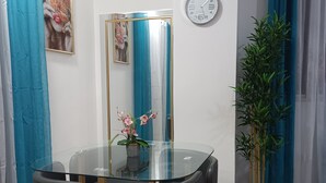 Uma área de refeições com mesa de vidro, duas cadeiras, um vaso de planta, um relógio e um espelho.