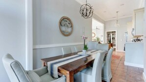 A dining area with a long wooden table, cushioned chairs, a hanging light fixture, and a framed artwork on the wall. - The Retreat on Broad @ Trustees' Garden (Savannah)