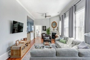 A spacious living room with a grey sofa, a coffee table, and a fireplace. There is a television mounted on the wall and a ceiling fan. - The Retreat on Broad @ Trustees' Garden (Savannah)