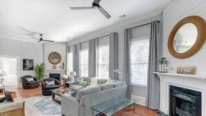 A living room with a grey sofa, a glass coffee table, and a fireplace. There are two ceiling fans, a large mirror, and a rug on the floor. - The Retreat on Broad @ Trustees' Garden (Savannah)