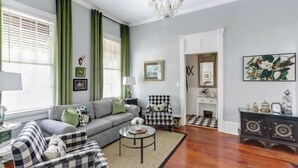 A living room with a grey sofa, checkered armchairs, a round coffee table, and a chandelier. - The Cottage @ Trustees' Garden | Walk To River! (Savannah)