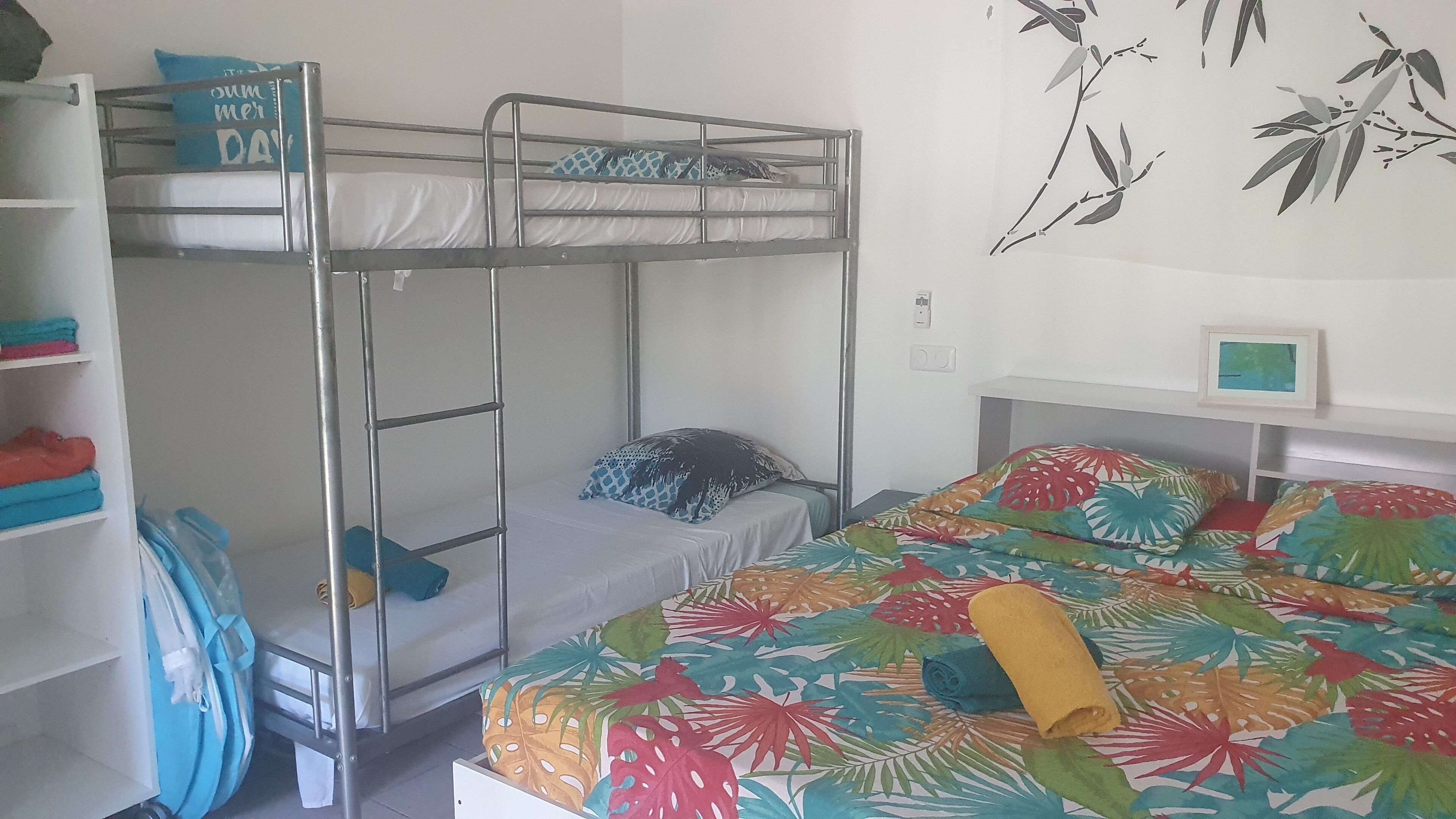 Camera con letto a castello, un letto inferiore, uno scaffale con vestiti piegati, uno zaino blu e un letto con motivo a foglie tropicali.
