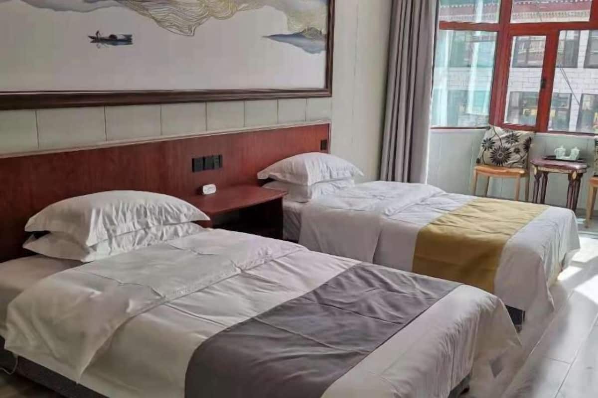 Ein Hotelzimmer mit zwei Betten, einem Holz-Kopfteil, einem Gemälde, einem Fenster mit Vorhängen und einem kleinen Tisch mit einem Stuhl.