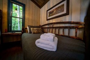 Une pièce confortable avec un lit, une fenêtre donnant sur les arbres, un tableau encadré au mur et une tête de lit en bois.